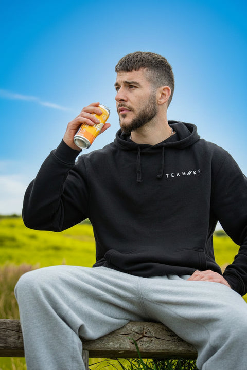 Man in black hoodie with 'TEAMWEAR' logo sitting outdoors, drinking from a can.