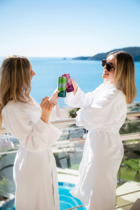 Two women in white robes holding drinks by a pool with a scenic background